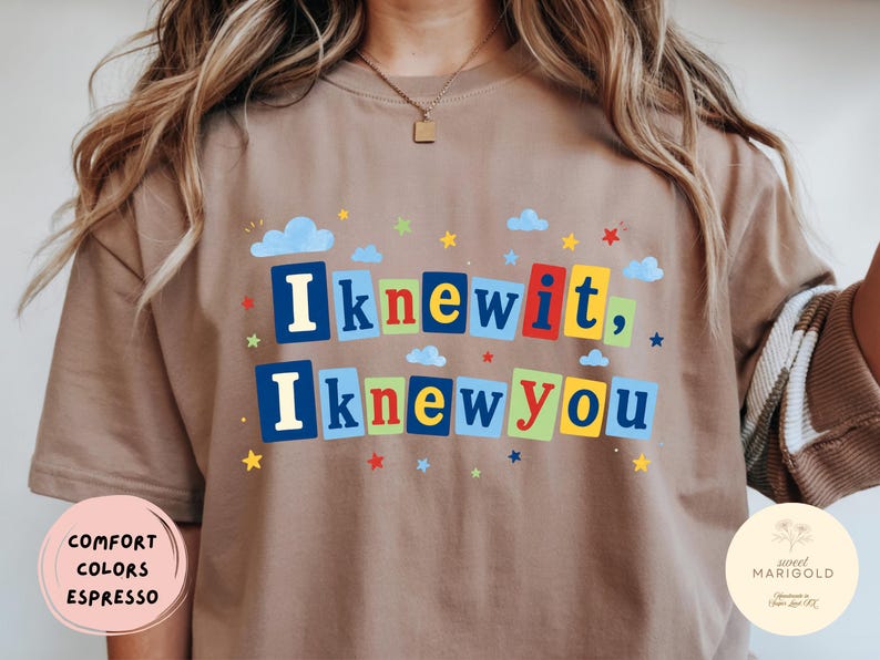 Op de afbeelding: Een bruin t-shirt met de tekst "Iknewit, Iknewyou" in kleurrijke blokletters. Het ontwerp is versierd met kleine blauwe wolken en diverse veelkleurige sterren die rond de tekst zijn verspreid, wat zorgt voor een speelse en levendige grafische uitstraling.