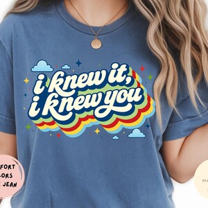 May include: A blue jean colored t-shirt featuring the white text I knew it, i knew you in a stacked, wavy font. The text is layered over a multi-colored rainbow graphic with red, yellow, and green stripes, accented by small blue clouds and stars.