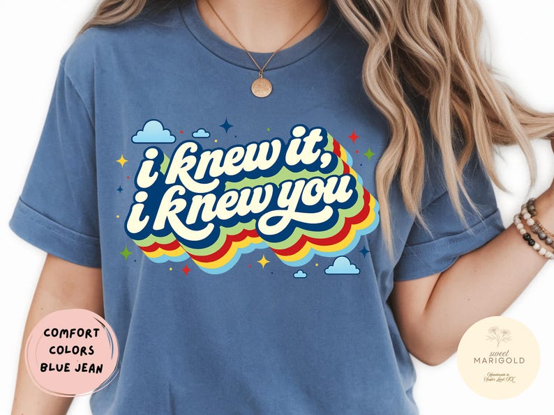 May include: A blue jean colored t-shirt featuring the white text I knew it, i knew you in a stacked, wavy font. The text is layered over a multi-colored rainbow graphic with red, yellow, and green stripes, accented by small blue clouds and stars.