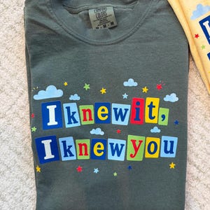 Op de afbeelding: Een donker olijfgroen t-shirt met ronde hals en de tekst "I knew it, I knew you" in kleurrijke blokletters. Het ontwerp is versierd met kleine blauwe wolken en veelkleurige sterren die rond de tekst verspreid zijn. Het shirt heeft een Comfort Colors label.