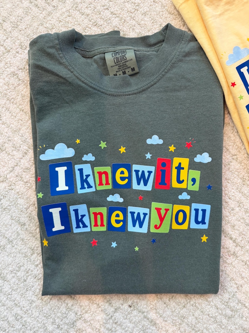 Op de afbeelding: Een donker olijfgroen t-shirt met ronde hals en de tekst "I knew it, I knew you" in kleurrijke blokletters. Het ontwerp is versierd met kleine blauwe wolken en veelkleurige sterren die rond de tekst verspreid zijn. Het shirt heeft een Comfort Colors label.