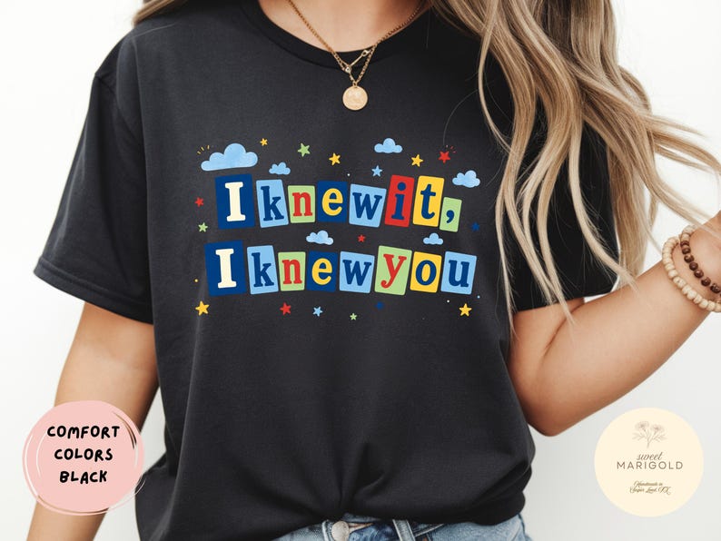 Op de afbeelding: Een zwart t-shirt met korte mouwen en een grafische print met de tekst "I knew it, I knew you" in kleurrijke blokletters. Het ontwerp bevat kleine blauwe wolken en veelkleurige sterren verspreid rond de tekst op de voorkant.