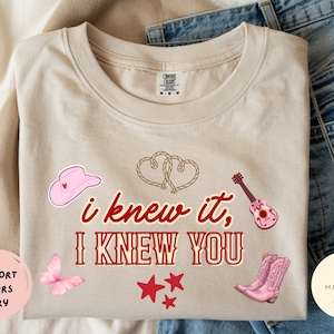 May include: An ivory t-shirt featuring the red text "i knew it, I KNEW YOU" surrounded by graphics of a pink cowboy hat, two rope hearts, a pink acoustic guitar, a pink butterfly, three red stars, and a pair of pink cowboy boots.