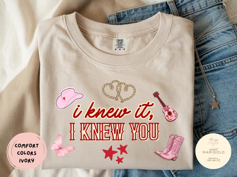 May include: An ivory t-shirt featuring the red text "i knew it, I KNEW YOU" surrounded by graphics of a pink cowboy hat, two rope hearts, a pink acoustic guitar, a pink butterfly, three red stars, and a pair of pink cowboy boots.