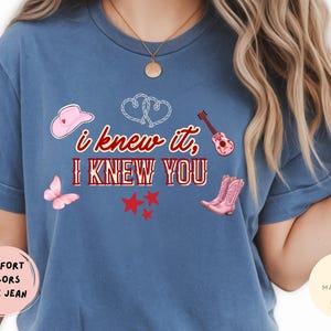 May include: A blue jean colored t-shirt featuring the text "i knew it, I KNEW YOU" in red and white. The design includes a pink cowboy hat, a pink acoustic guitar, a pink butterfly, three red stars, a pair of pink cowboy boots, and two white hearts.