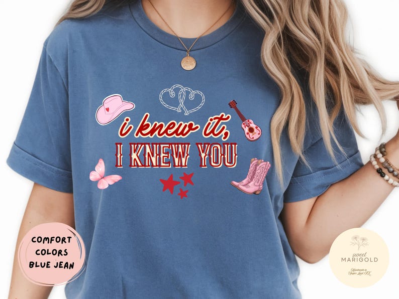 May include: A blue jean colored t-shirt featuring the text "i knew it, I KNEW YOU" in red and white. The design includes a pink cowboy hat, a pink acoustic guitar, a pink butterfly, three red stars, a pair of pink cowboy boots, and two white hearts.