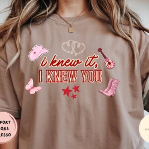 May include: An espresso-colored t-shirt featuring the red text "i knew it, I KNEW YOU" centered on the chest. The design includes a pink cowboy hat, a pink butterfly, a small guitar, a pair of pink cowboy boots, red stars, and two white hearts.