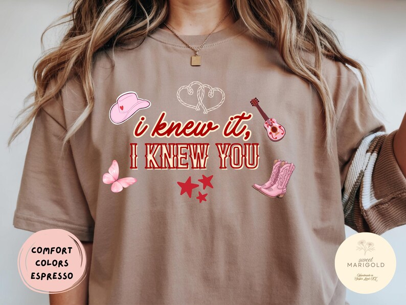 May include: An espresso-colored t-shirt featuring the red text "i knew it, I KNEW YOU" centered on the chest. The design includes a pink cowboy hat, a pink butterfly, a small guitar, a pair of pink cowboy boots, red stars, and two white hearts.