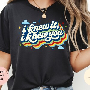 May include: A black t-shirt featuring the text "i knew it, i knew you" in a white, stylized font. The text is layered over a colorful, wavy graphic design in shades of blue, green, yellow, and red, accented with small blue clouds and yellow stars.