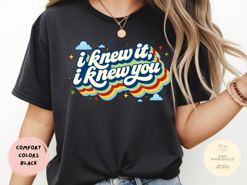 May include: A black t-shirt featuring the text "i knew it, i knew you" in a white, stylized font. The text is layered over a colorful, wavy graphic design in shades of blue, green, yellow, and red, accented with small blue clouds and yellow stars.