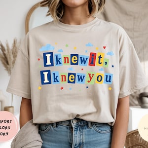 Op de afbeelding: Een ivoorkleurig t-shirt met korte mouwen en de tekst "Iknewit, Iknewyou" in kleurrijke blokletters. De letters staan op individuele rechthoekige achtergronden in blauw, rood, geel en groen, geaccentueerd door kleine wolkjes en sterretjes.