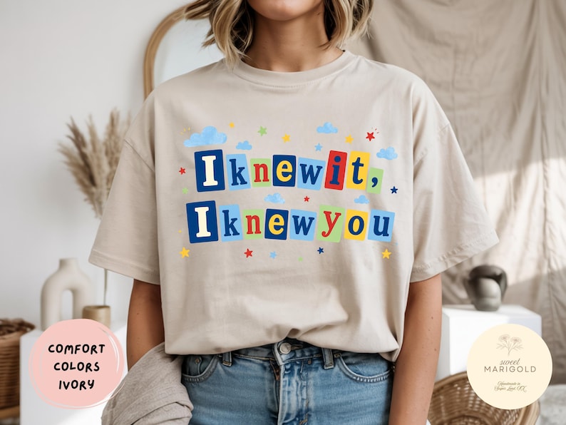Op de afbeelding: Een ivoorkleurig t-shirt met korte mouwen en de tekst "Iknewit, Iknewyou" in kleurrijke blokletters. De letters staan op individuele rechthoekige achtergronden in blauw, rood, geel en groen, geaccentueerd door kleine wolkjes en sterretjes.