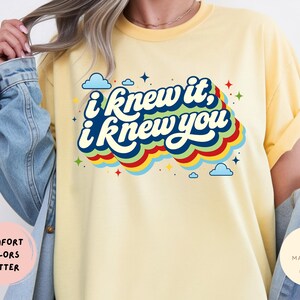 May include: A butter yellow t-shirt featuring the dark blue text I knew it, I knew you in a retro font. The design includes colorful layered waves in red, yellow, green, and blue, accented with small blue clouds and multi-colored star shapes.