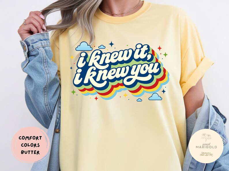May include: A butter yellow t-shirt featuring the dark blue text I knew it, I knew you in a retro font. The design includes colorful layered waves in red, yellow, green, and blue, accented with small blue clouds and multi-colored star shapes.