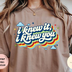 May include: An espresso-colored t-shirt featuring the text "i knew it, i knew you" in dark blue script. The text is layered over a multi-colored wavy graphic in light blue, green, yellow, and red, accented by small blue clouds and colorful stars.