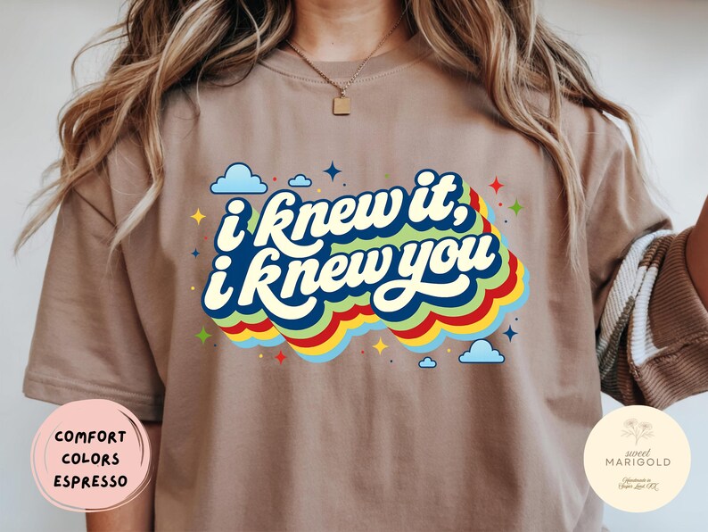 May include: An espresso-colored t-shirt featuring the text "i knew it, i knew you" in dark blue script. The text is layered over a multi-colored wavy graphic in light blue, green, yellow, and red, accented by small blue clouds and colorful stars.