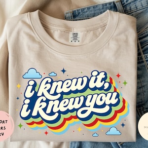 May include: An ivory-colored t-shirt featuring a graphic print with the text "i knew it, i knew you" in a bold, dark blue font. The text is layered over a multi-colored, wavy rainbow design in green, yellow, and red, accented by small blue clouds and stars.