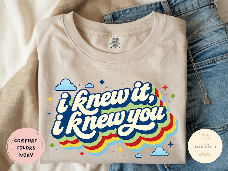 May include: An ivory-colored t-shirt featuring a graphic print with the text "i knew it, i knew you" in a bold, dark blue font. The text is layered over a multi-colored, wavy rainbow design in green, yellow, and red, accented by small blue clouds and stars.
