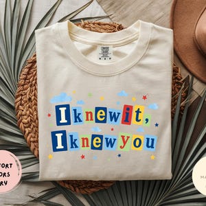 Op de afbeelding: Een ivoorkleurig t-shirt met korte mouwen en de tekst "Iknewit, Iknewyou" in een kleurrijke blokletterstijl. Het ontwerp bevat kleine blauwe wolken en rode, gele en blauwe sterren die rondom de tekst op de voorkant van het shirt zijn verspreid.
