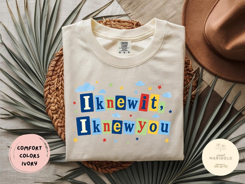 Op de afbeelding: Een ivoorkleurig t-shirt met korte mouwen en de tekst "Iknewit, Iknewyou" in een kleurrijke blokletterstijl. Het ontwerp bevat kleine blauwe wolken en rode, gele en blauwe sterren die rondom de tekst op de voorkant van het shirt zijn verspreid.