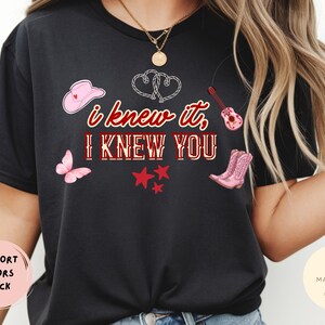 May include: A black t-shirt featuring the text "I KNEW IT, I KNEW YOU" in red and white lettering. The design includes a pink cowboy hat, a pink butterfly, a pink acoustic guitar, pink cowboy boots, three red stars, and two white rope hearts.