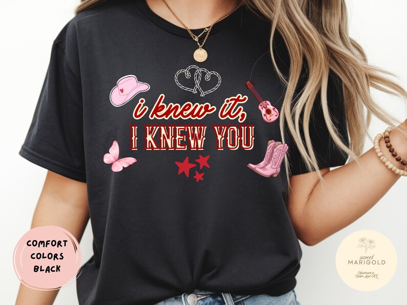 May include: A black t-shirt featuring the text "I KNEW IT, I KNEW YOU" in red and white lettering. The design includes a pink cowboy hat, a pink butterfly, a pink acoustic guitar, pink cowboy boots, three red stars, and two white rope hearts.