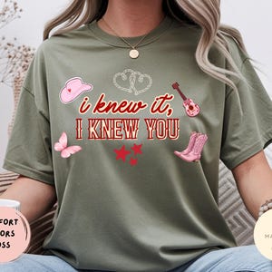 May include: An olive green short-sleeve t-shirt featuring the red text "i knew it, I KNEW YOU" surrounded by graphic illustrations of a pink cowboy hat, a pink acoustic guitar, a pink butterfly, three red stars, and two white outlined hearts.