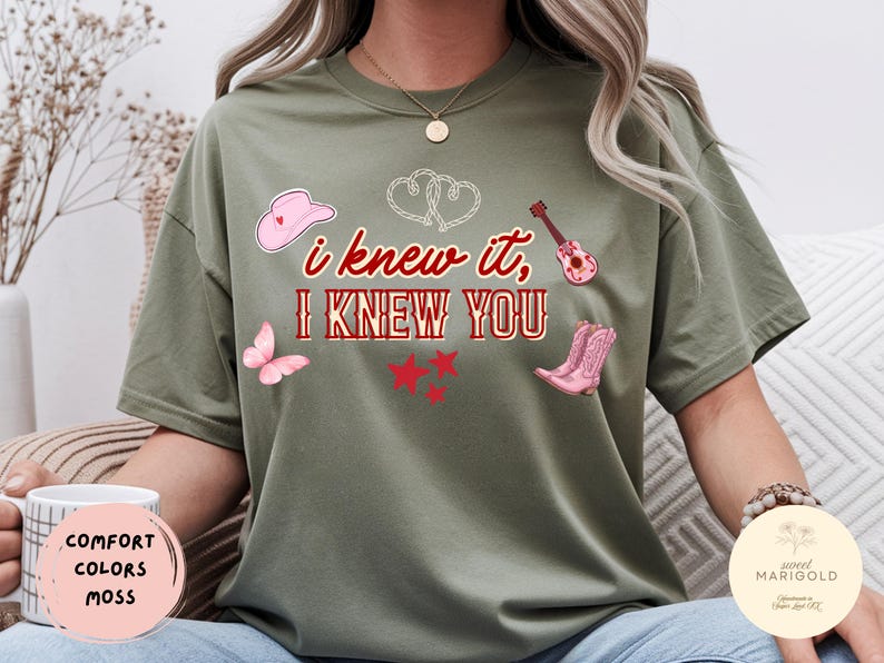 May include: An olive green short-sleeve t-shirt featuring the red text "i knew it, I KNEW YOU" surrounded by graphic illustrations of a pink cowboy hat, a pink acoustic guitar, a pink butterfly, three red stars, and two white outlined hearts.