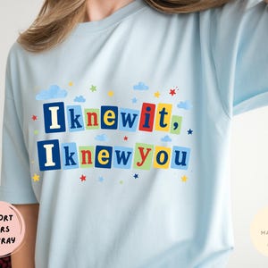 Op de afbeelding: Een lichtblauw t-shirt met de tekst "Iknewit, Iknewyou" in kleurrijke blokletters. Het ontwerp bevat kleine blauwe wolken en veelkleurige sterren verspreid rond de tekst, die op rechthoekige achtergronden is geplaatst.
