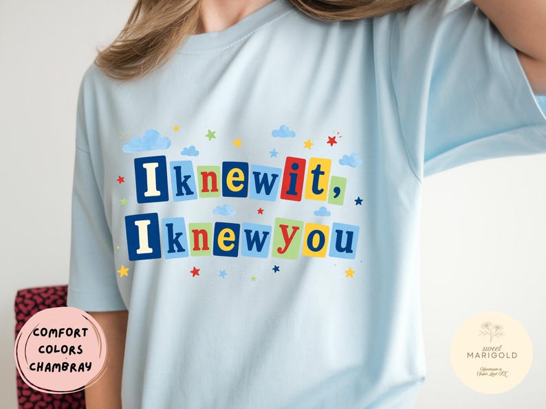 Op de afbeelding: Een lichtblauw t-shirt met de tekst "Iknewit, Iknewyou" in kleurrijke blokletters. Het ontwerp bevat kleine blauwe wolken en veelkleurige sterren verspreid rond de tekst, die op rechthoekige achtergronden is geplaatst.