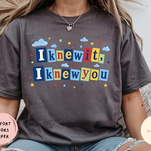 Op de afbeelding: Een antracietgrijs T-shirt met korte mouwen en de tekst "I knew it, I knew you" in kleurrijke blokletters. Het ontwerp bevat kleine blauwe wolken en veelkleurige sterren verspreid rond de tekst, wat zorgt voor een speelse grafische print.