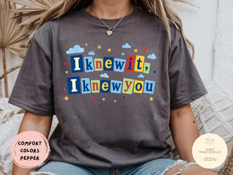 Op de afbeelding: Een antracietgrijs T-shirt met korte mouwen en de tekst "I knew it, I knew you" in kleurrijke blokletters. Het ontwerp bevat kleine blauwe wolken en veelkleurige sterren verspreid rond de tekst, wat zorgt voor een speelse grafische print.