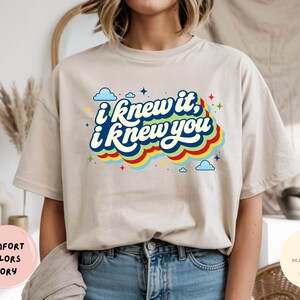 May include: An ivory short-sleeve t-shirt featuring a graphic print with the text "i knew it, i knew you" in white script. The text is layered over a series of colorful, wavy horizontal stripes in red, yellow, green, and blue, accented by small blue clouds and multi-colored stars.