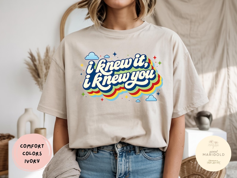 May include: An ivory short-sleeve t-shirt featuring a graphic print with the text "i knew it, i knew you" in white script. The text is layered over a series of colorful, wavy horizontal stripes in red, yellow, green, and blue, accented by small blue clouds and multi-colored stars.