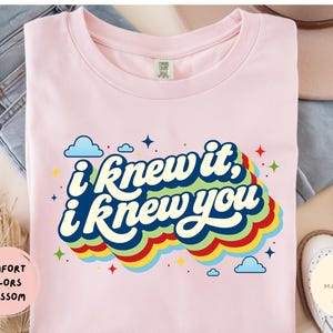 May include: A light pink t-shirt featuring the dark blue text "i knew it, i knew you" in a retro font. The text is layered over a multi-colored, wavy rainbow graphic with small blue clouds and colorful star accents scattered around the design.
