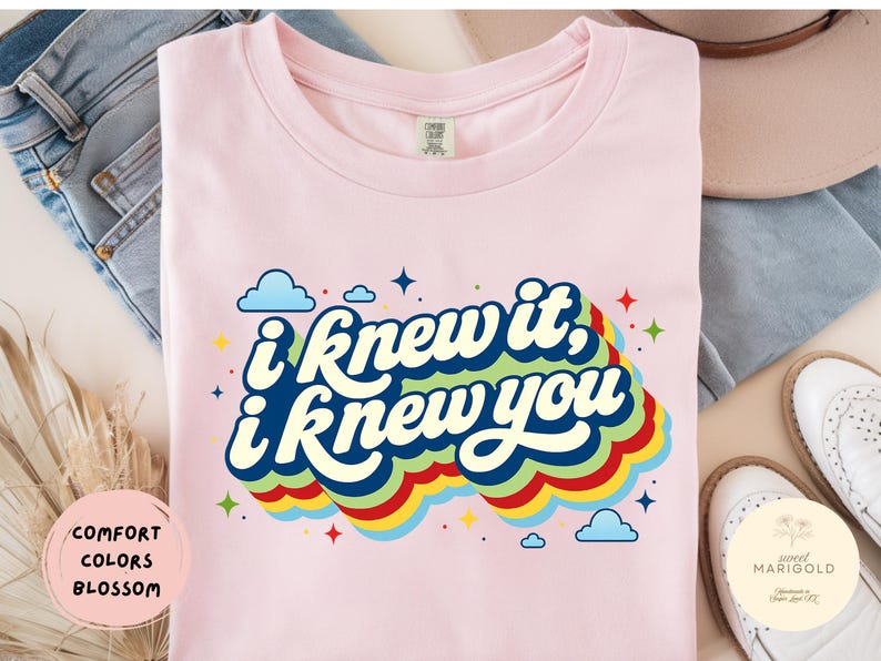 May include: A light pink t-shirt featuring the dark blue text "i knew it, i knew you" in a retro font. The text is layered over a multi-colored, wavy rainbow graphic with small blue clouds and colorful star accents scattered around the design.