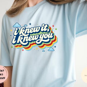 May include: A light blue t-shirt featuring a graphic print with the text "i knew it, i knew you" in white letters. The text is layered over a multi-colored, wavy, retro-style background with small blue clouds and colorful star accents.
