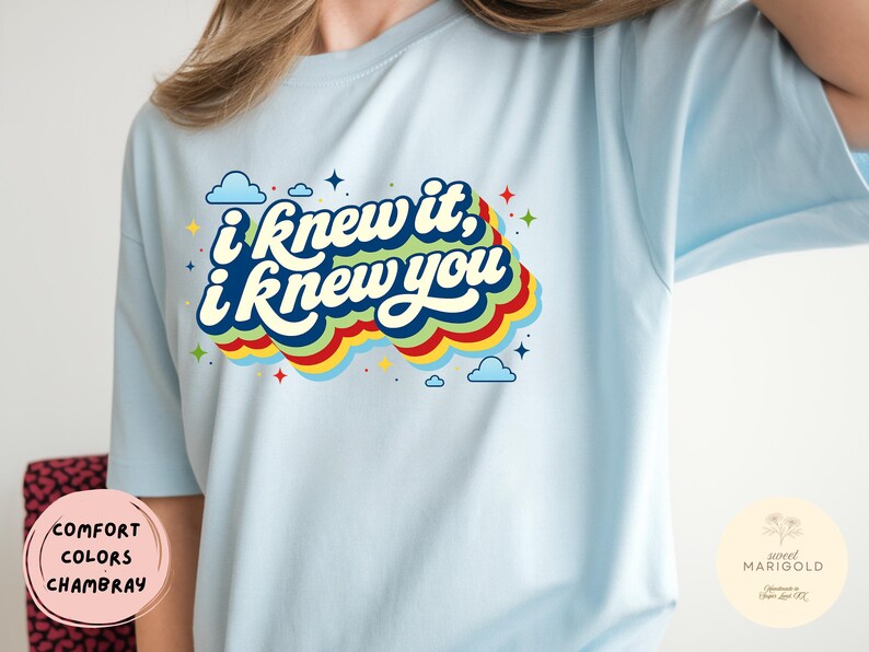 May include: A light blue t-shirt featuring a graphic print with the text "i knew it, i knew you" in white letters. The text is layered over a multi-colored, wavy, retro-style background with small blue clouds and colorful star accents.