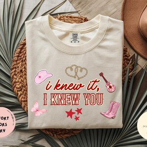 May include: An ivory short-sleeve t-shirt featuring the red text "i knew it, I KNEW YOU" centered on the chest. The design includes a pink cowboy hat, a pink acoustic guitar, pink cowboy boots, a pink butterfly, three red stars, and two rope hearts.