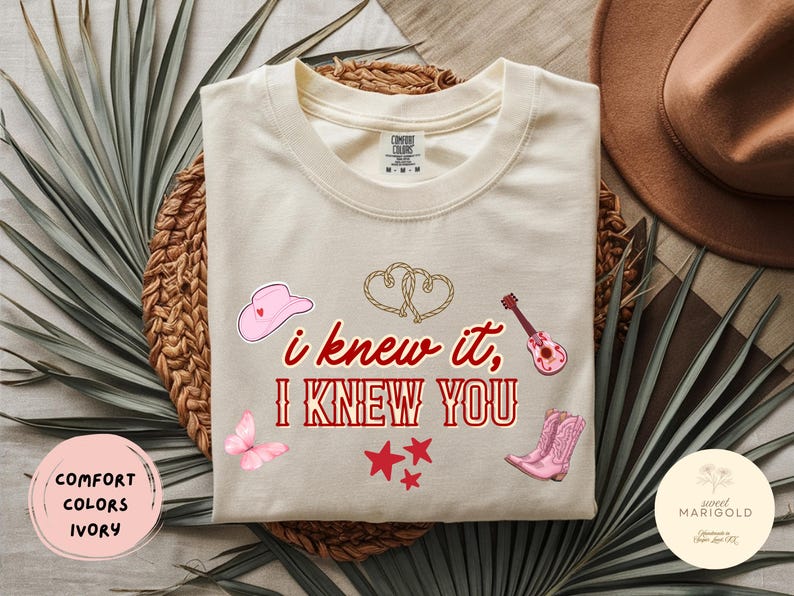 May include: An ivory short-sleeve t-shirt featuring the red text "i knew it, I KNEW YOU" centered on the chest. The design includes a pink cowboy hat, a pink acoustic guitar, pink cowboy boots, a pink butterfly, three red stars, and two rope hearts.