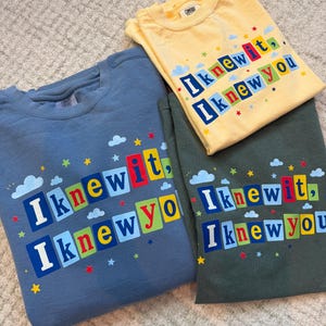 Op de afbeelding: Drie T-shirts met ronde hals in blauw, geel en donkergroen. Elk shirt is voorzien van de tekst "I knew it, I knew you" in kleurrijke blokletters, omringd door kleine geprinte wolkjes en sterretjes. De shirts hebben een casual ontwerp.