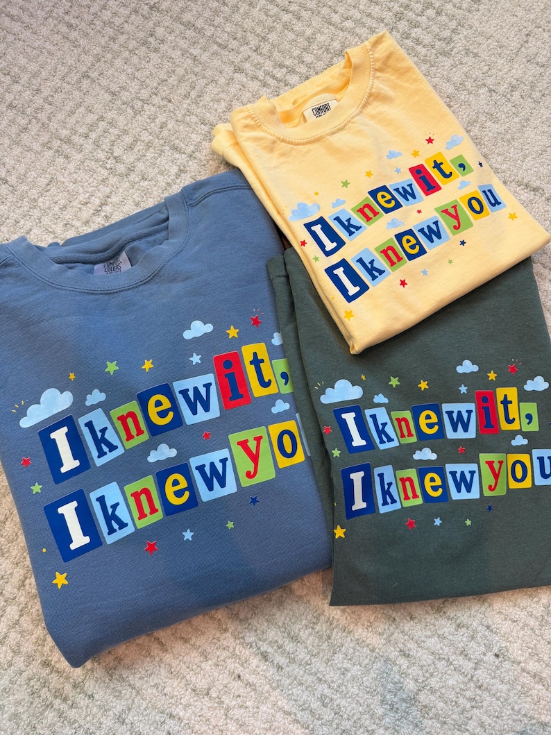 Op de afbeelding: Drie T-shirts met ronde hals in blauw, geel en donkergroen. Elk shirt is voorzien van de tekst "I knew it, I knew you" in kleurrijke blokletters, omringd door kleine geprinte wolkjes en sterretjes. De shirts hebben een casual ontwerp.