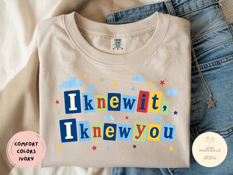 Op de afbeelding: Een ivoorkleurig t-shirt met ronde hals en de tekst "Iknewit, Iknewyou" in kleurrijke blokletters. Het ontwerp is versierd met kleine blauwe wolkjes en veelkleurige sterretjes die rondom de tekst zijn verspreid. Comfort Colors label aanwezig.