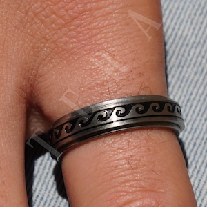 Może przedstawiać: Srebrny pierścionek z czarnym wzorem fal. Pierścionek jest noszony na palcu.