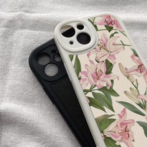 May include: Two phone cases, one white with a floral design and one black with a camera lens cutout. The white case has a pink and green floral pattern with lilies.