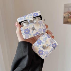 May include: A white and purple floral patterned case for AirPods. The case has a flap that closes with a button.