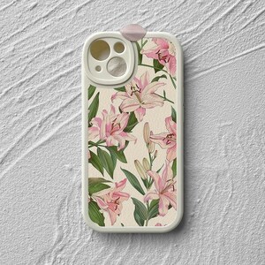 May include: White phone case with a floral design featuring pink lilies. The case has a clear camera lens cover with a ring around it.