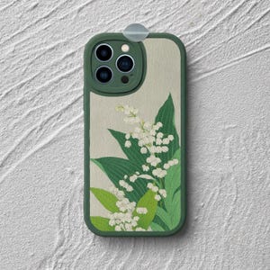 May include: A dark green smartphone case featuring a light beige background with a botanical illustration of white lily of the valley flowers and green leaves. The case has a raised camera cutout with three circular lenses and a flash.
