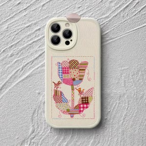 Peut inclure: Une coque de téléphone couleur crème ornée d'une illustration centrale en style patchwork représentant une grande fleur. La fleur est composée de divers motifs de tissus colorés, incluant carreaux, vichy et pois, avec deux petits chats sur la tige.