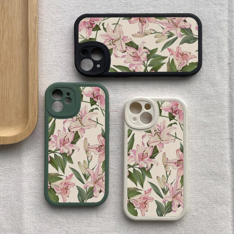 May include: Three phone cases with a floral design. The cases are black, green, and white. The floral design features pink lilies on a cream background.